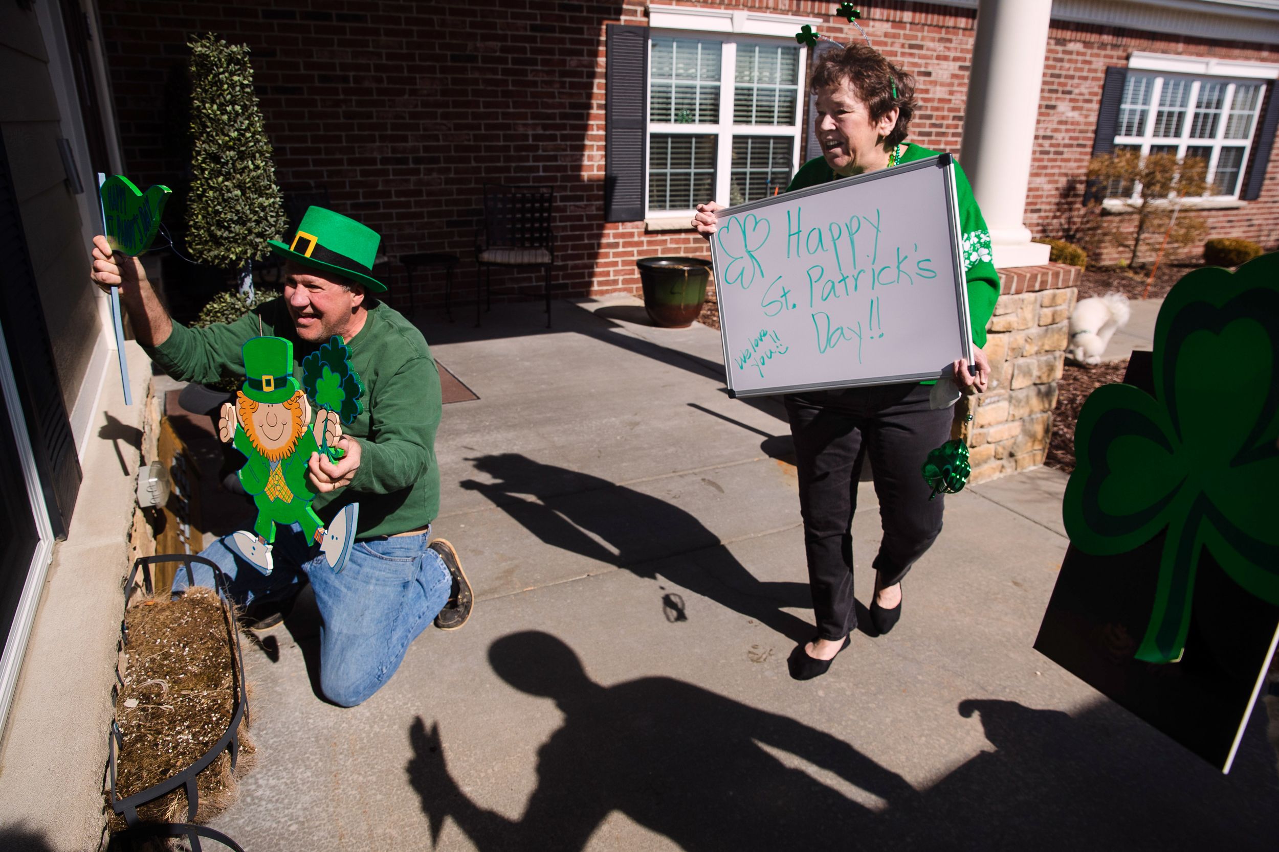 Two adults in St. Patrick’s day attire hold signs figures celebrating the day outside the window of a building for residents inside to see.