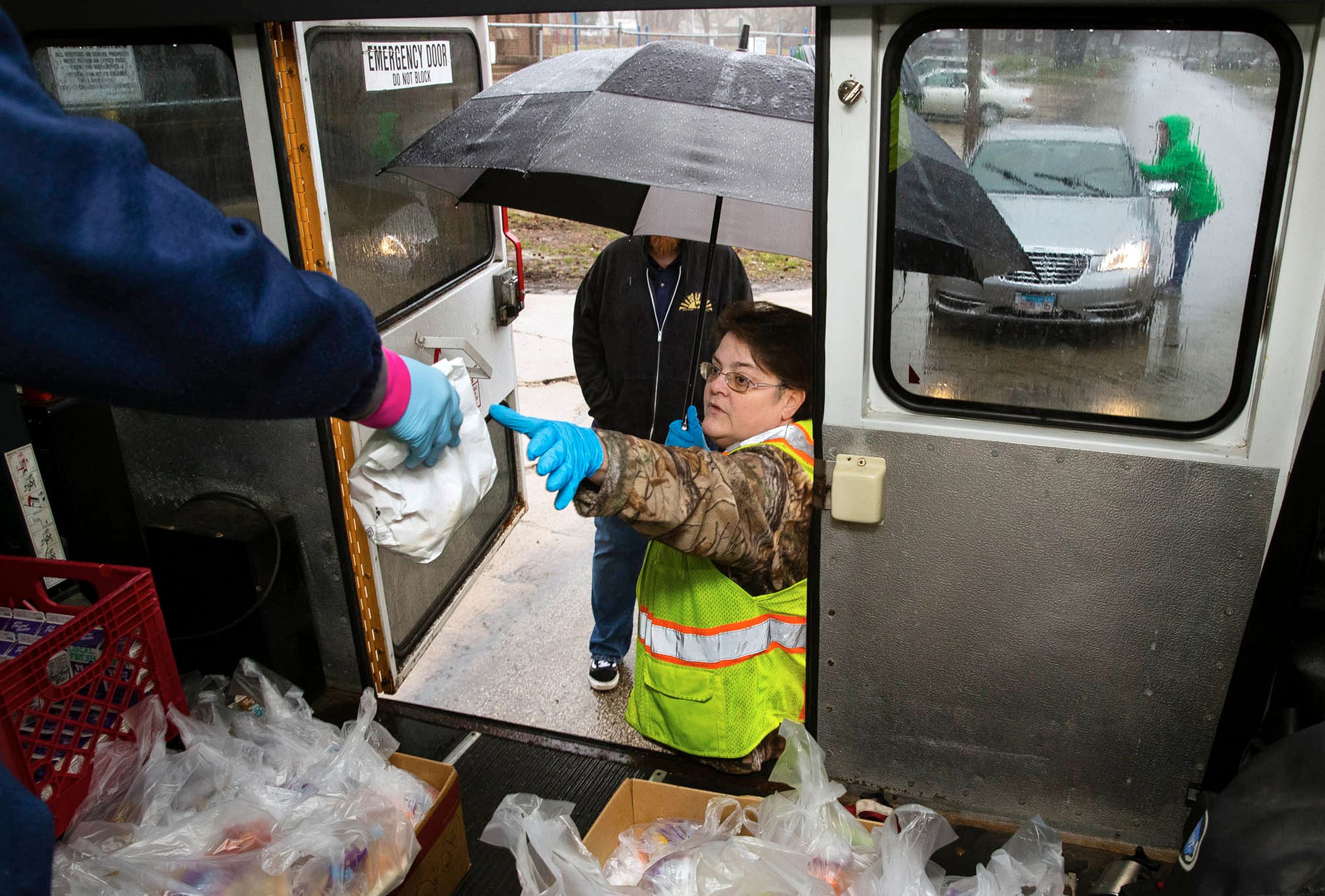 A volunteer holding an umbrella reaches into the back door of a school bus to receive a bagged meal to distribute to waiting cars at a school meal distribution event during COVID-19 closure at Feitshans Elementary, Springfield, Illinois.