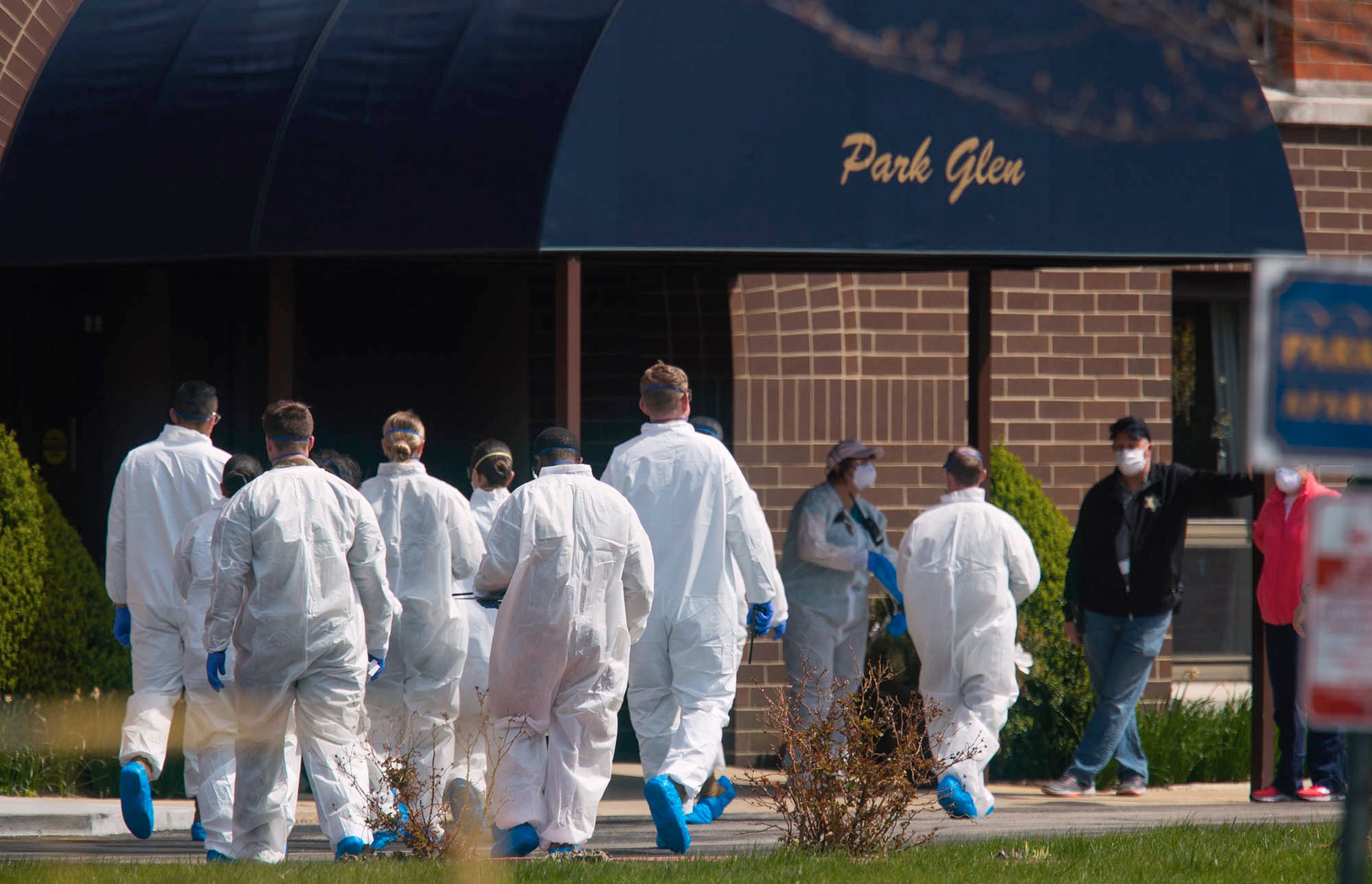 A group of workers clad in white protective suits with blue gloves and booties approach the entrance to a building.