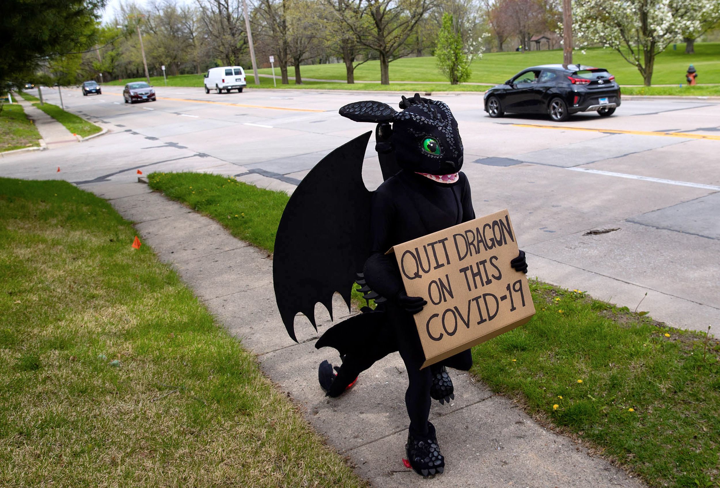 Man dressed as cartoon dragon holds sign saying, “QUIT DRAGON THIS COVID-19” as he walks along sidewalk near four lane street.