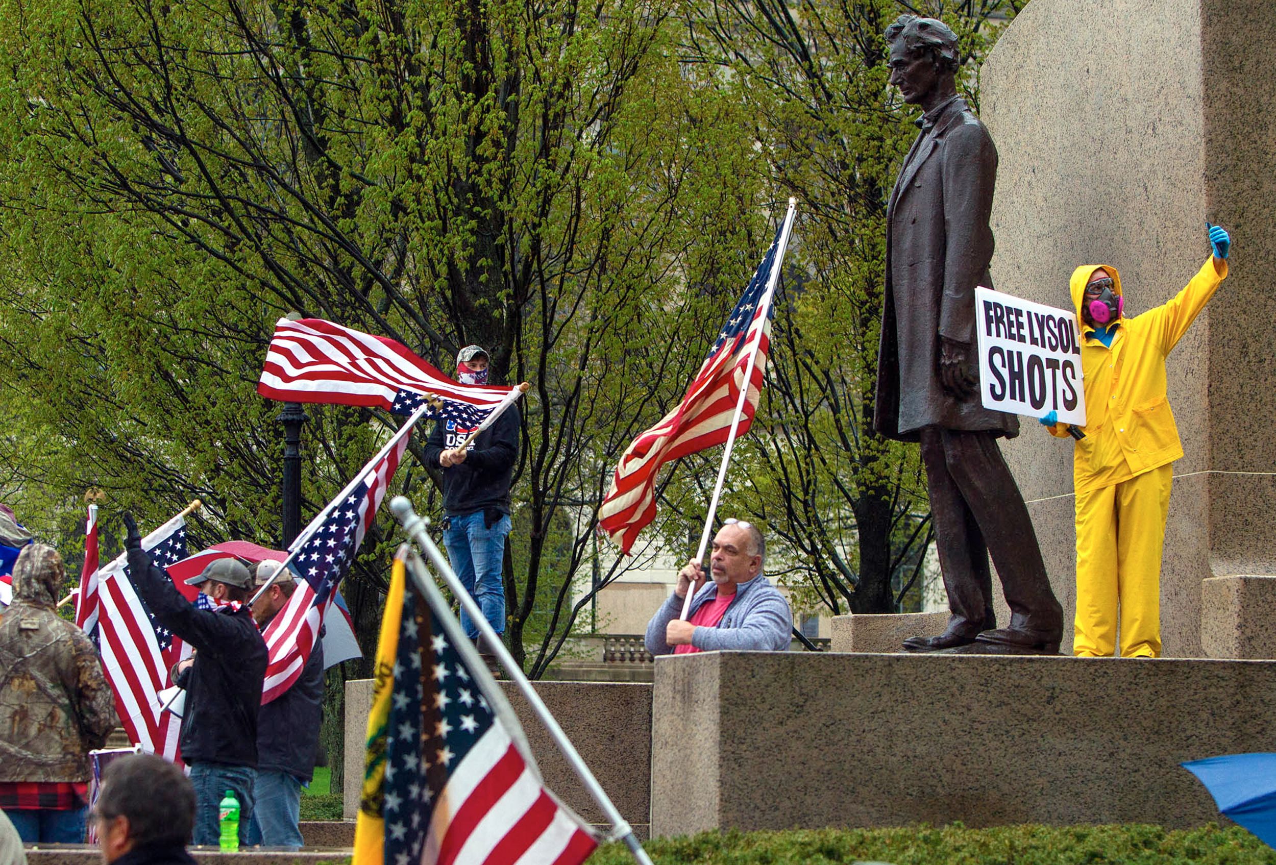 A man dressed in yellow rain gear and a full respirator holds a satirical sign mocking Trump's disinfectant comments at protest against Illinois’ stay-at-home order at the Illinois Capitol, April 2020.