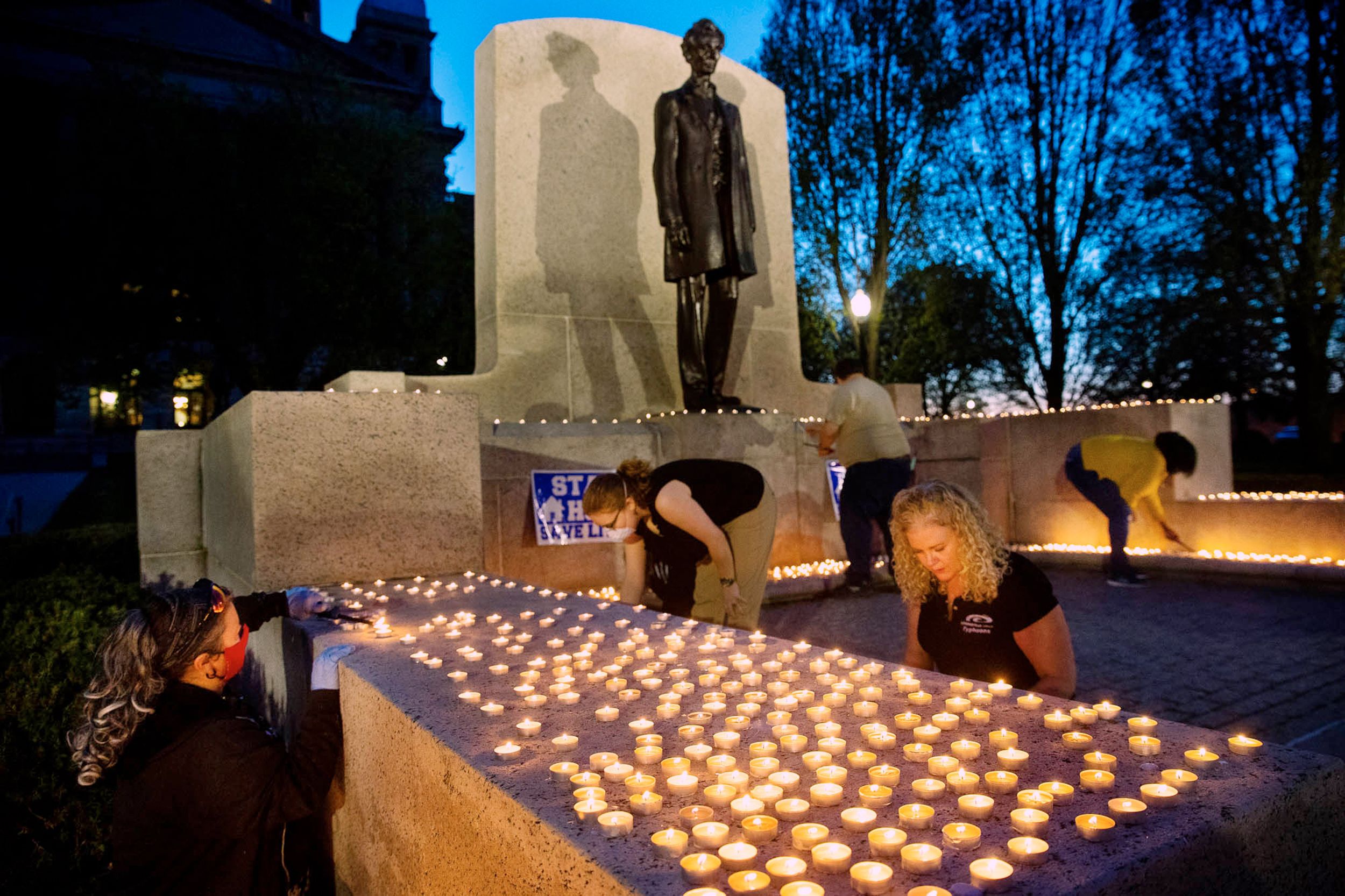 In the waning moments of daylight, five volunteers arrange and light candles around Abraham Lincoln statue at Illinois Capitol to honor 1,933 COVID-19 deaths.