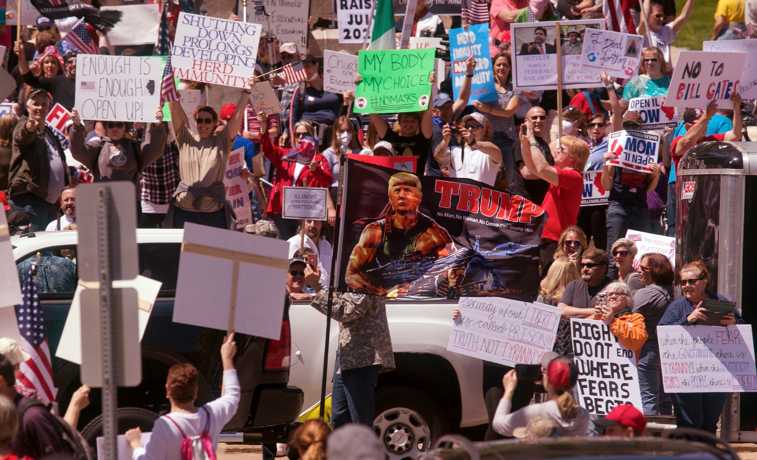 A flag of a Rambo-esque Donald Trump holding a machine gun waves in front of a crowd protesters opposing stay-at-home order at Illinois Capitol, Springfield, May 2020.