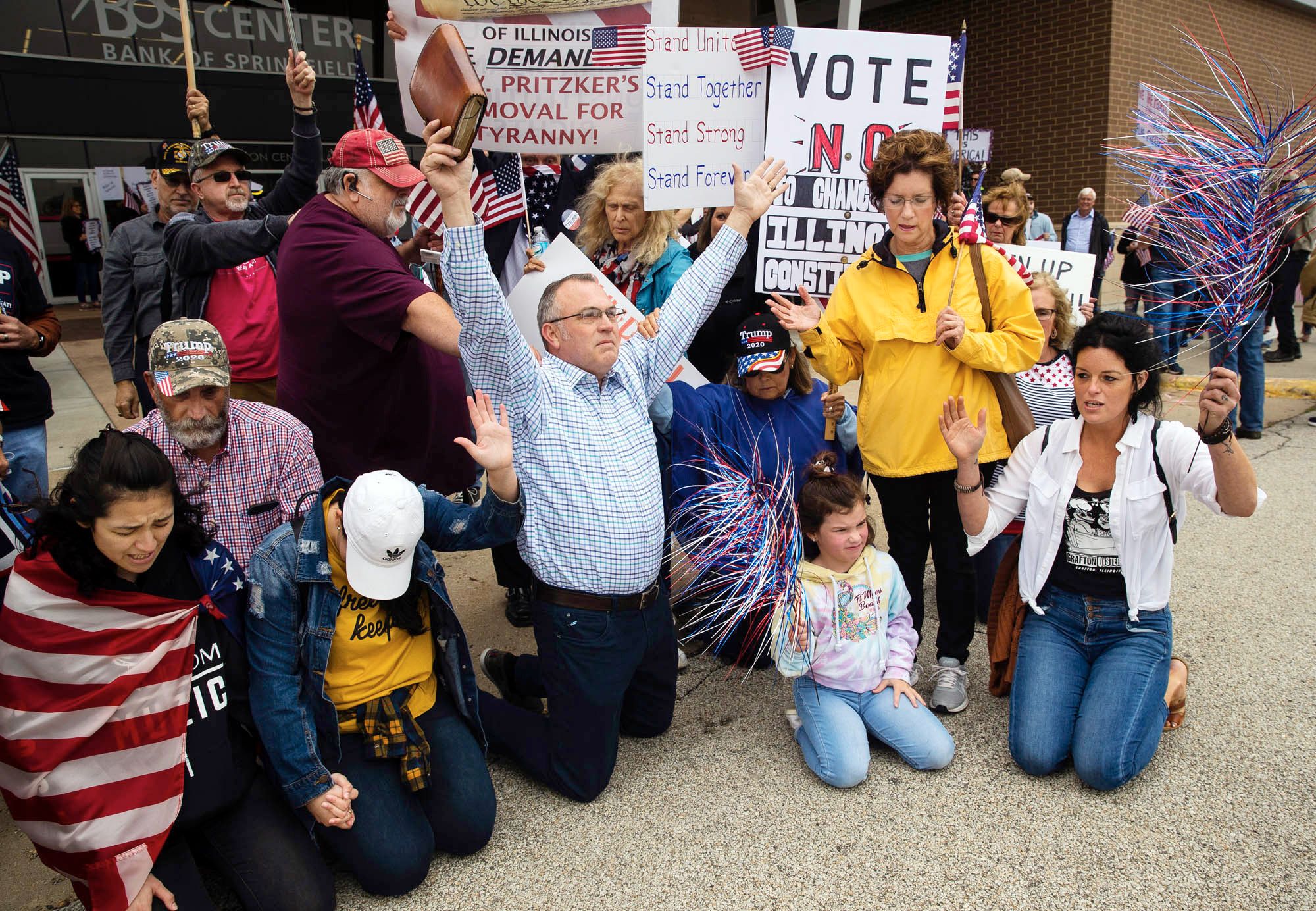 Surrounded by a small, praying crowd, a man opens his arms wide while holding a Bible during 'Open Illinois' protest outside the Bank of Springfield Center, May 2020.