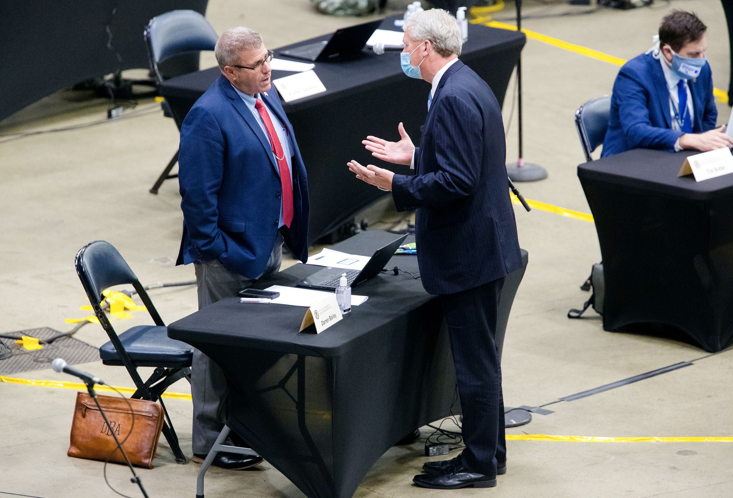 Two standing men, one wearing a mask, one not, confer at a table.