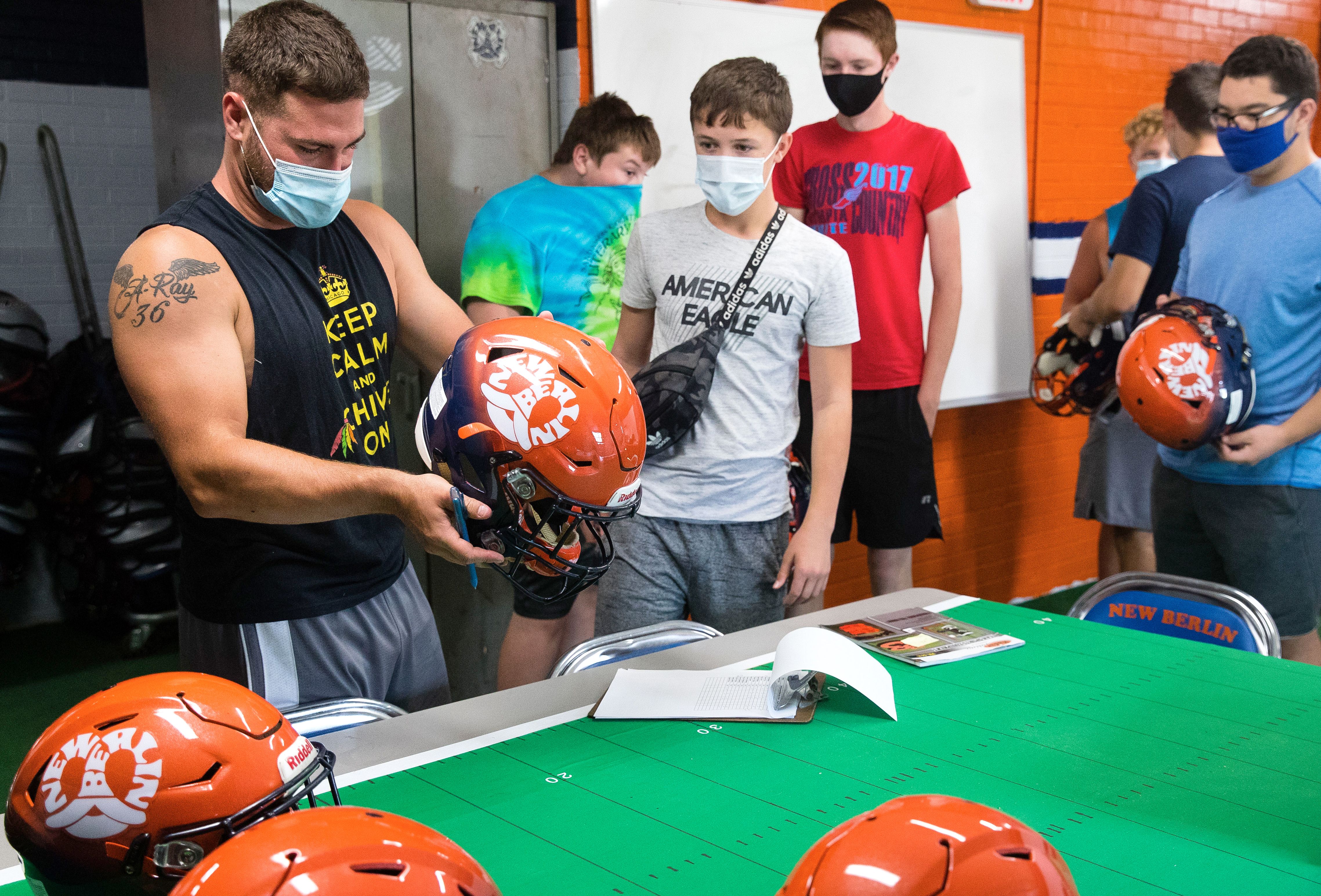 An assistant football coach collects team helmets during COVID-19 practice restrictions, July 2020. All are wearing face masks.