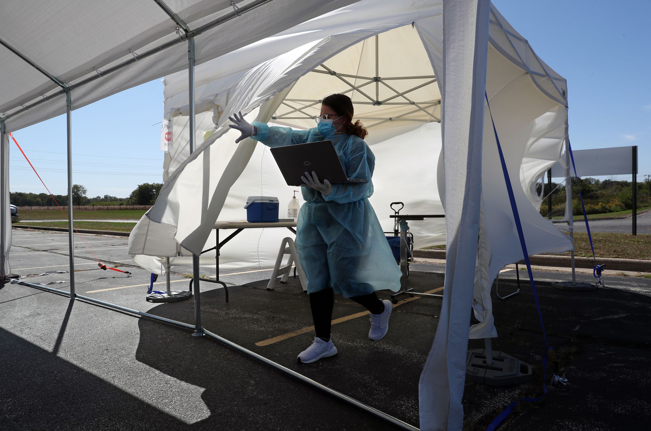 Healthcare worker in blue gown and mask holding at laptop steps out of a tent at a parking lot COVID-19 test station.