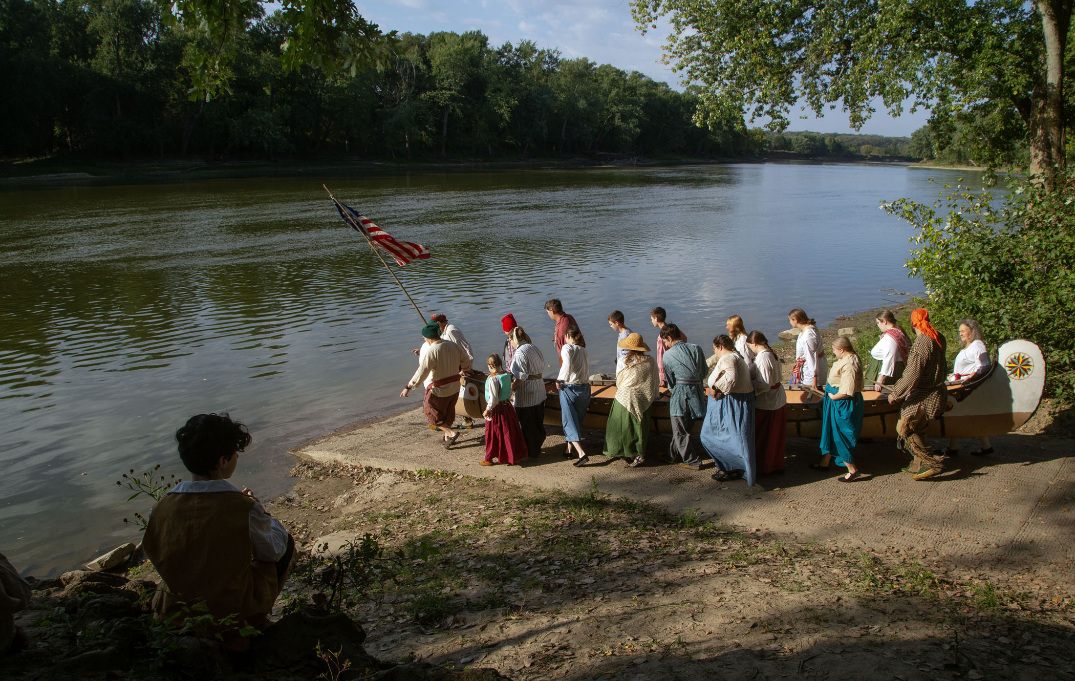 Reenactors in colonial-era dress carry a canoe to the Wabash River as a young boy watches from the bank above.